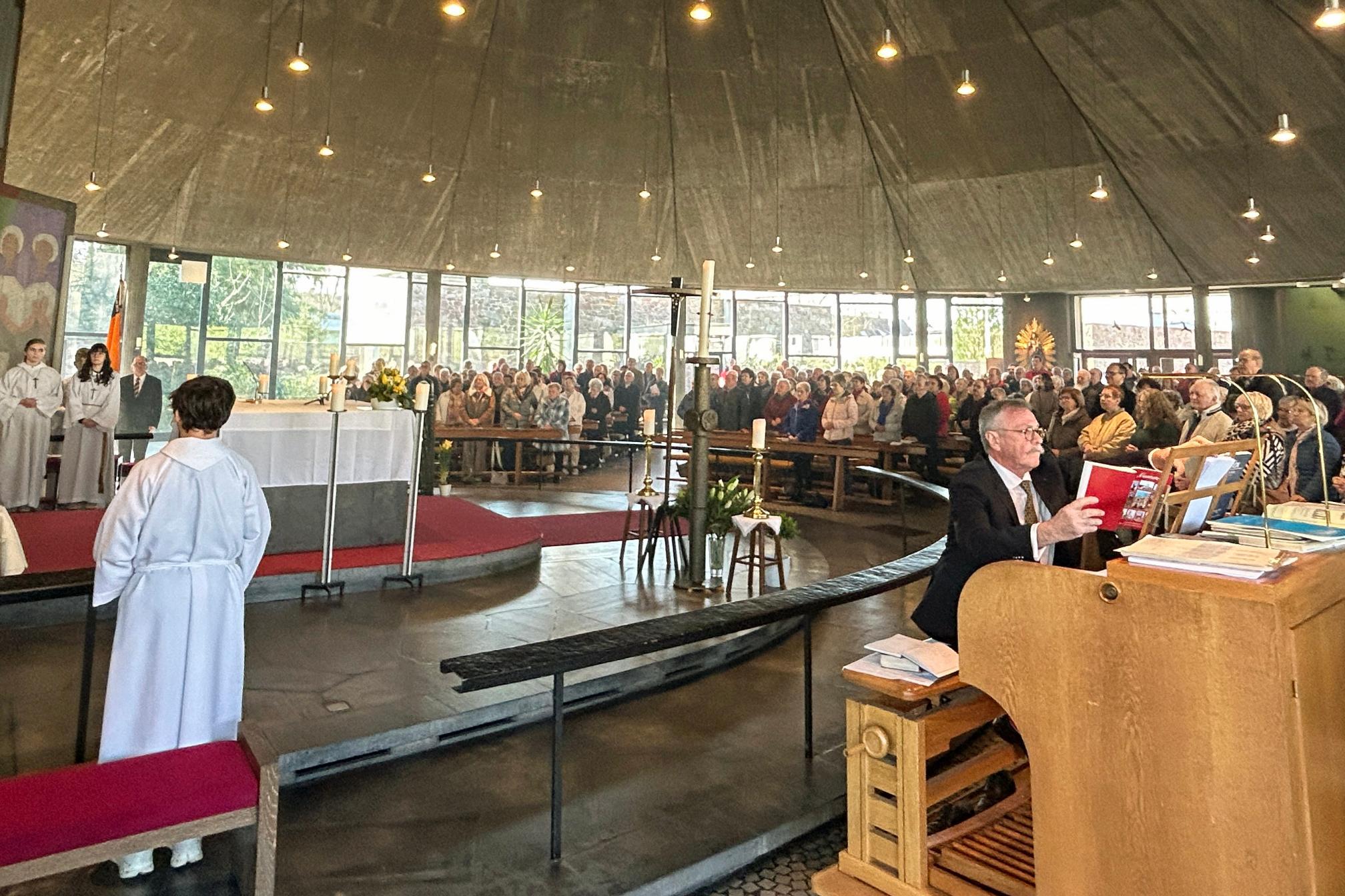 Innenaufnahme einer großen, modern gestalteten Kirche während eines festlichen Gottesdienstes. Die Gemeinde füllt den gesamten Raum, viele Personen sitzen oder stehen dicht beieinander in den Bankreihen, die halbkreisförmig um den Altar angeordnet sind. Der Altar ist zentral positioniert, weiß gedeckt und von liturgischen Diensten umgeben, darunter mehrere Personen in hellen liturgischen Gewändern. Im linken Bereich stehen weitere Personen im Altarraum und beteiligen sich am Gottesdienst. Rechts im Vordergrund sitzt eine Person an einer großen Orgel und spielt, vor sich Noten und Bücher auf dem Instrument. Der Kirchenraum ist lichtdurchflutet durch hohe Fenster im Hintergrund, und zahlreiche kleine Lampen hängen von der dunklen, zeltartigen Decke herab. Insgesamt vermittelt das Bild den Eindruck eines feierlichen und gut besuchten Festhochamts.